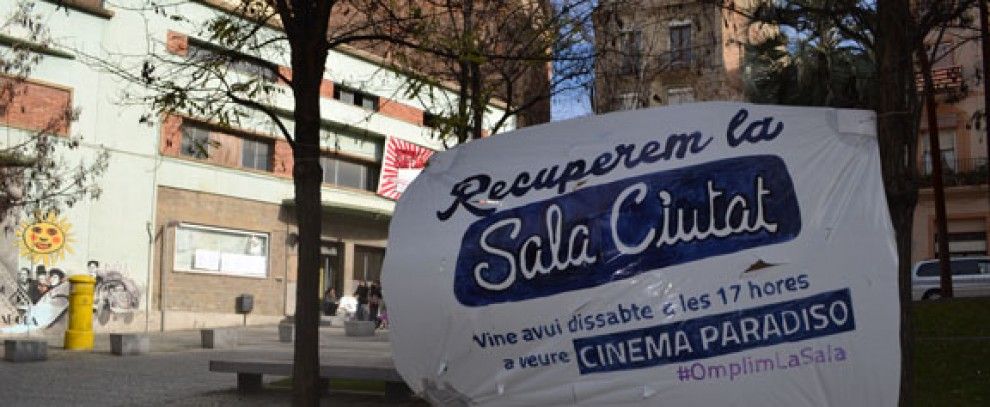 Pancarta de 'Recuperem la Sala Ciutat' a la plaça Sant Ignasi. Al fons, joves a la porta de l'equipament
