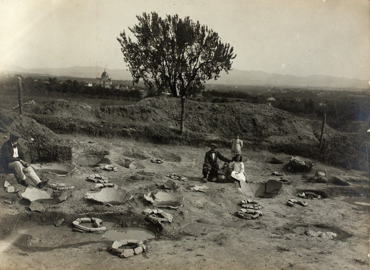 Vista del camp de dolies de la vil·la romana de la Salut, al fons, Joan Vila Cinca, (dret) i Amadeu Aragay (assegut) acompanyats d'un nen i una nena sense identificar (1912 - 1913)