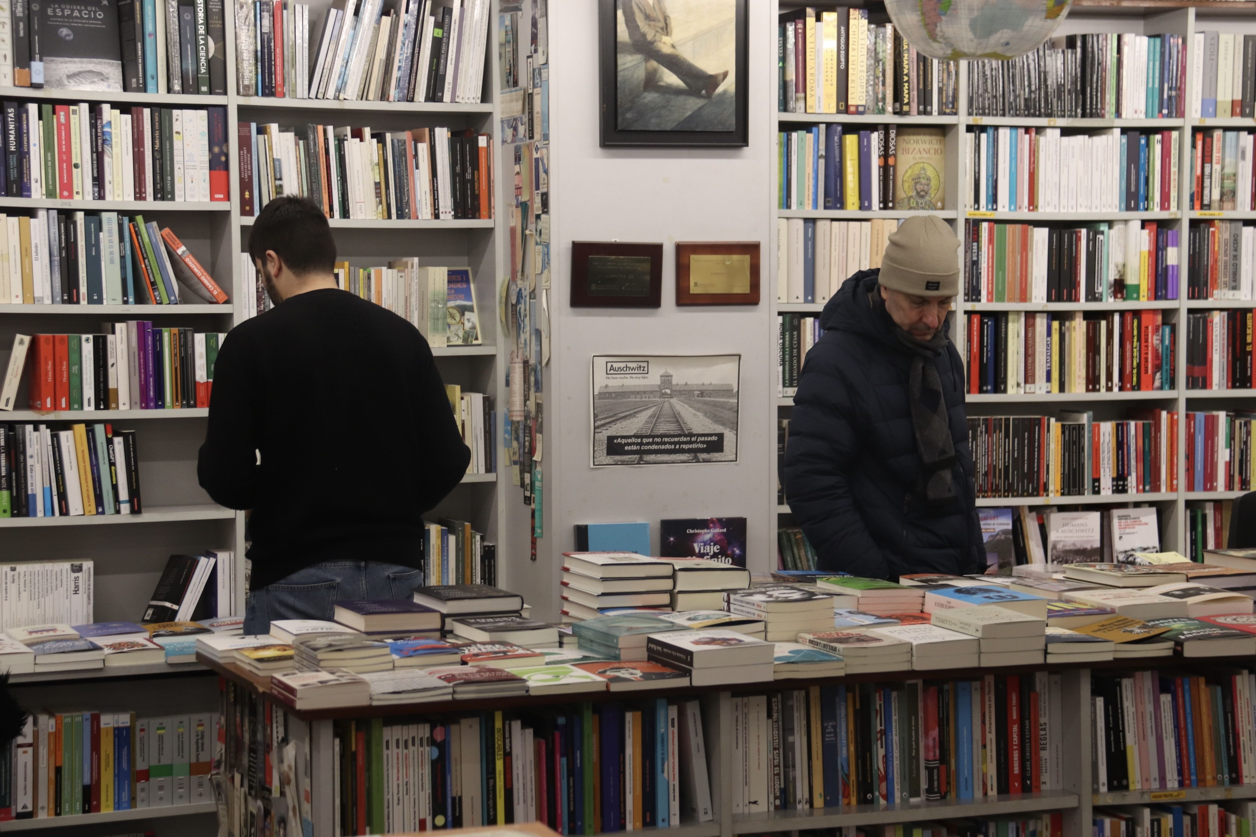 Un llibreter de la 22 de Girona col·loca llibres a les lleixes mentre un client mira alguns exemplars que hi ha sobre una de les taules